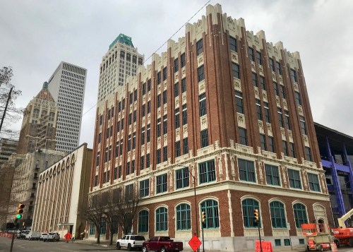 Historic brick building with ornate details, surrounded by modern skyscrapers under a cloudy sky. Construction work nearby.