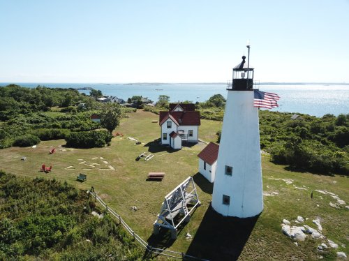 A scenic lighthouse with an American flag, surrounded by greenery and a view of the ocean on a clear day.