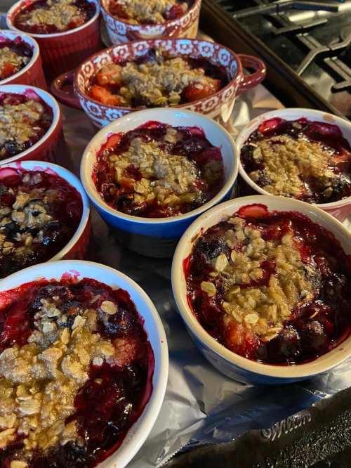 Baked fruit desserts in small dishes, topped with a crumbly oat topping, arranged on a baking tray.