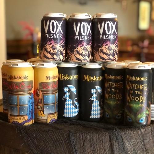 A display of craft beer cans, featuring "Vox Pilsner" and "Miskatonic" brews like "Duffle Coat" and "Watcher in the Woods."