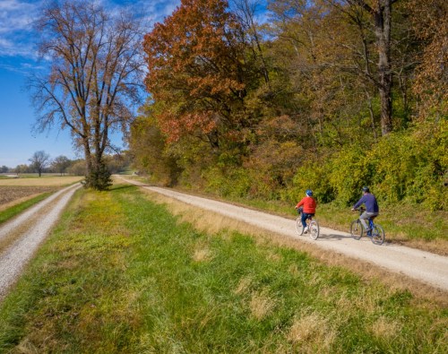 Two cyclists ride along a gravel path surrounded by trees with autumn foliage and open fields under a blue sky.