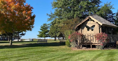A wooden gazebo with butterfly decor surrounded by green grass and colorful trees under a clear blue sky.