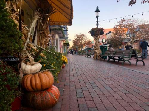 A charming brick pathway lined with autumn decorations, pumpkins, and people strolling in a quaint town.