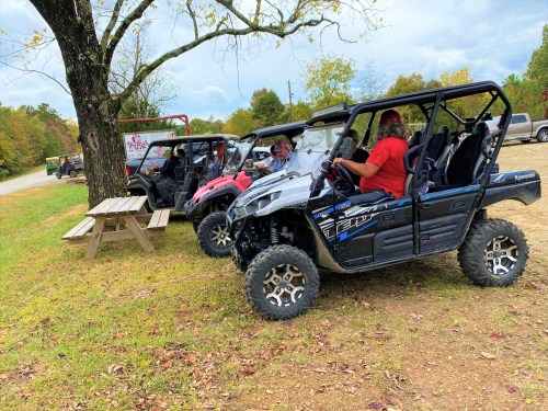 Four off-road vehicles parked near a picnic table under a tree, with a cloudy sky in the background.