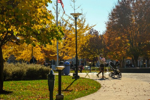 A sunny park scene with autumn foliage, people walking, and a parent with a stroller near green tables.
