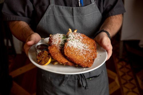 A person in an apron holds a plate with two breaded cutlets, fries, and a lemon wedge, garnished with cheese.
