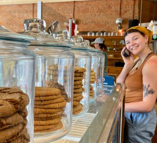 A smiling barista on the phone stands behind a counter with glass jars filled with cookies.