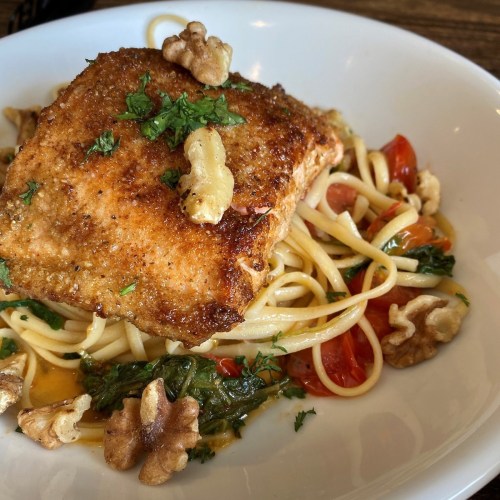 A plate of salmon on a bed of linguine, garnished with walnuts, cherry tomatoes, and fresh herbs.