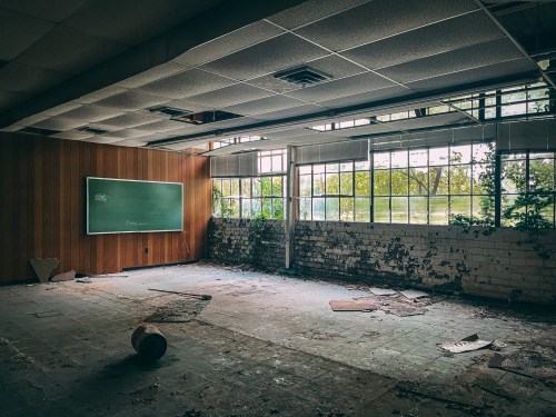 Abandoned classroom with a chalkboard, broken windows, and debris scattered on the floor, illuminated by natural light.