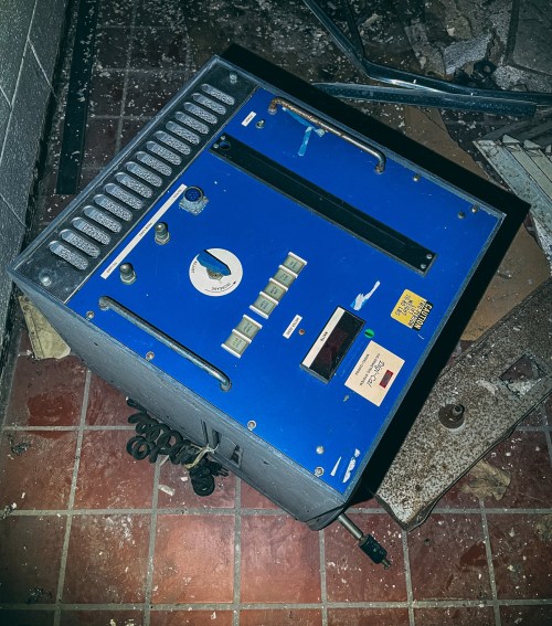 An old blue control panel with buttons and a dial, resting on a tiled floor amidst debris.