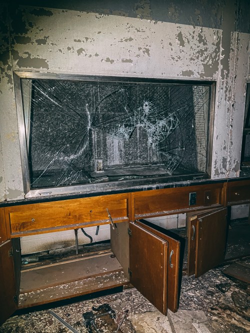 An abandoned room with a shattered glass display and open wooden cabinets, showing signs of neglect and decay.