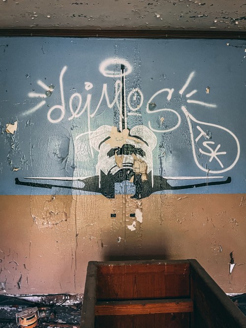 Graffiti on a peeling wall featuring the word "deimos" and a stylized design, with a wooden box in the foreground.
