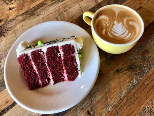 A slice of red velvet cake with frosting and sprinkles next to a yellow cup of latte art on a wooden table.