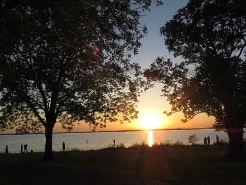 Sunset over a lake, framed by trees, with silhouettes of people walking along the shore.