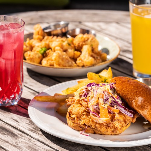 A plate with a fried chicken sandwich topped with slaw, fries, and two drinks, with a bowl of fried cauliflower in the background.