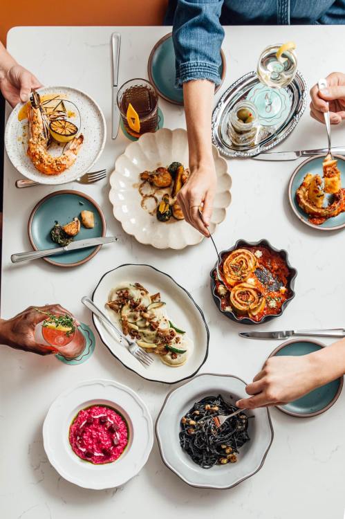 A table filled with various colorful dishes, hands reaching for food, and drinks in a vibrant dining setting.
