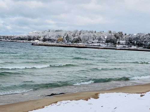 A snowy shoreline with gentle waves lapping at the beach, surrounded by trees and a distant pier.