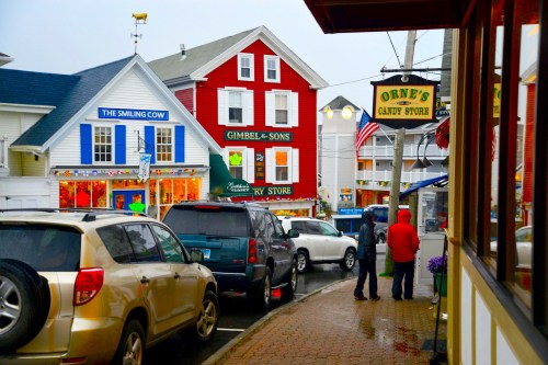 Colorful storefronts line a rainy street, with people walking and cars parked along the curb.