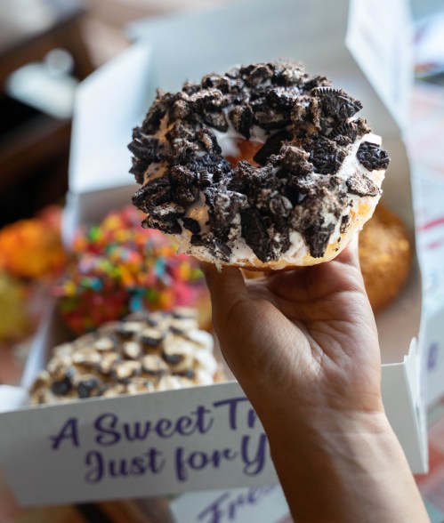 A hand holds a large donut topped with crushed cookies, with a colorful box of assorted donuts in the background.