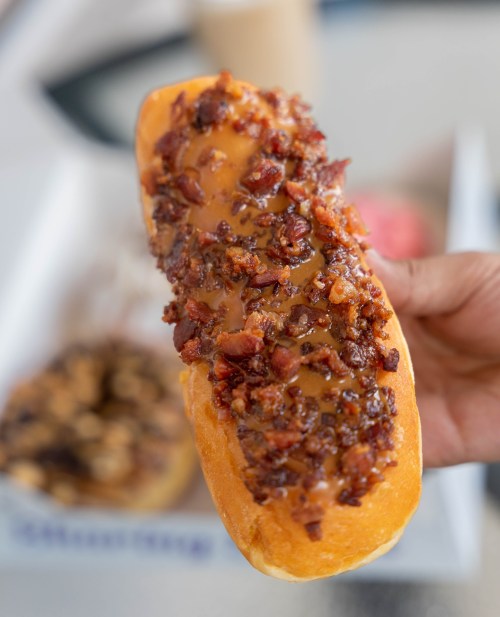A hand holding a glazed donut topped with crispy bacon bits, with other donuts in the background.
