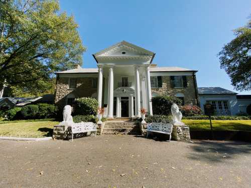 A large stone mansion with white columns, flanked by lion statues and benches, surrounded by greenery under a clear blue sky.