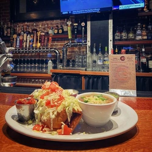 A plate of tacos topped with lettuce and tomatoes, served with salsa and a bowl of soup, in a bar setting.