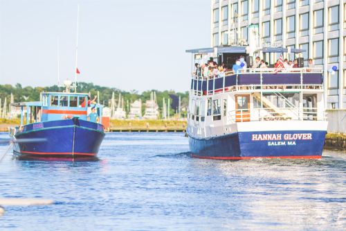 Two boats navigate a calm waterway, with a building and marina in the background on a sunny day.