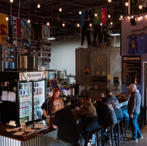 A lively brewery interior with patrons at the bar, colorful banners, and a variety of drinks displayed.