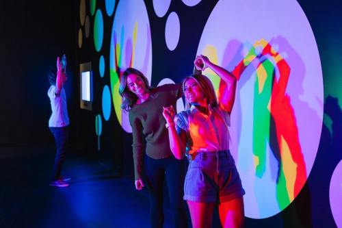 Two women pose playfully in front of colorful, abstract wall art, while a person dances in the background.