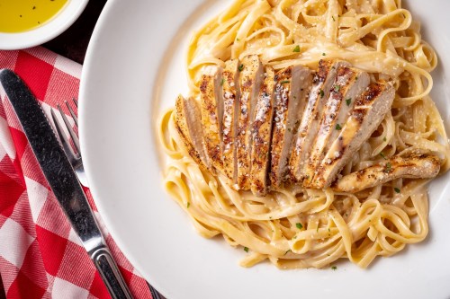 A plate of fettuccine alfredo topped with sliced grilled chicken, served with a checkered tablecloth and utensils.
