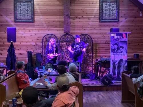 A duo performs on stage in a rustic venue, with an audience seated at tables enjoying the music.