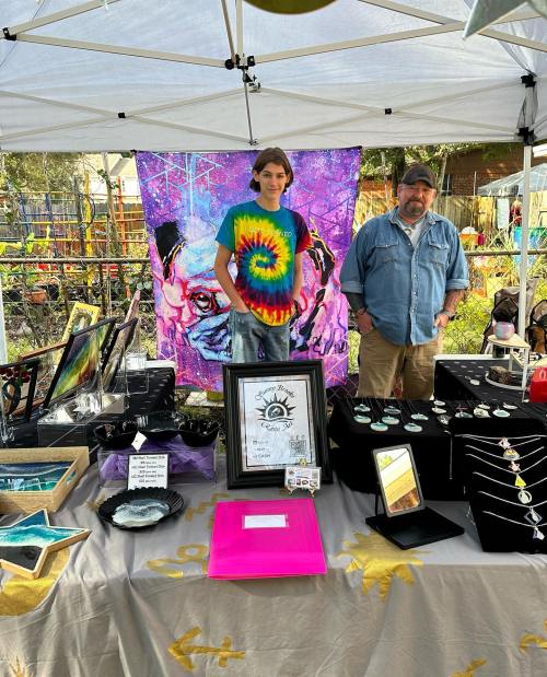 A young person in a tie-dye shirt and an older man stand behind a craft booth with various handmade items and a colorful backdrop.