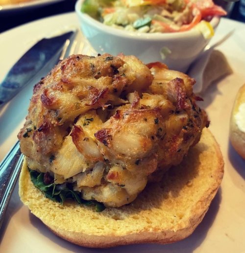 A close-up of a crab cake on a bun, garnished with lettuce, alongside a small bowl of salad.