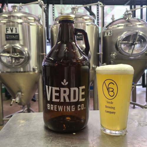 A growler and a pint glass of beer from Verde Brewing Co. in front of brewing tanks.