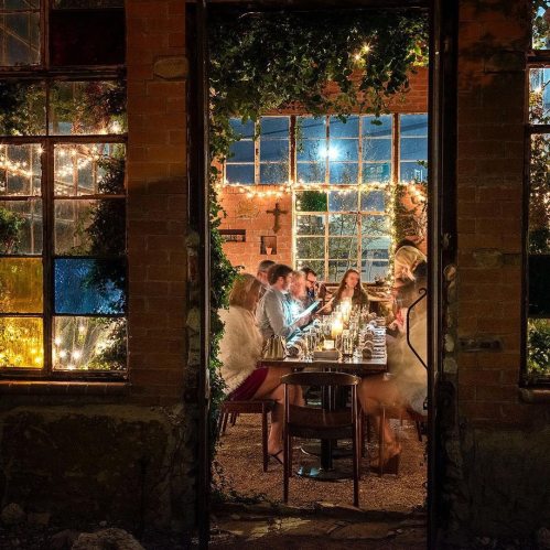 A cozy dinner scene through a window, with guests gathered around a table illuminated by warm lights and greenery.