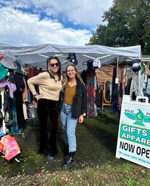 Two women pose together in front of a colorful outdoor market stall filled with clothing and accessories.
