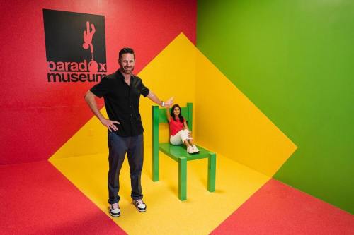A man stands beside a large green chair, while a woman sits on it, in a colorful room at the Paradox Museum.