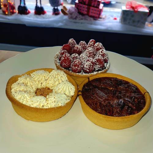 Three dessert tarts on a plate: one with whipped cream, one with raspberries and powdered sugar, and a chocolate tart.