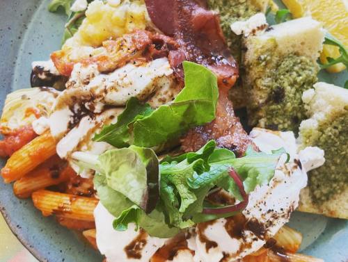A colorful plate of pasta with sauce, bacon, greens, and a side of bread and orange slices.