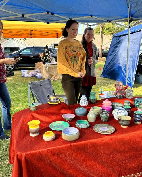 Two women stand behind a table of colorful pottery under a tent, with a child playing nearby.