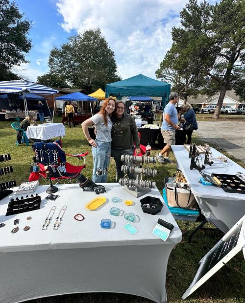 Two women pose together at a craft fair, surrounded by tables displaying jewelry and crafts under a blue sky.