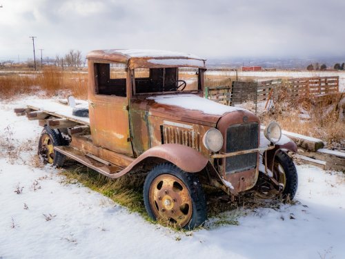 An old, rusty truck covered in snow, sitting in a field with tall grass and wooden pallets in the background.