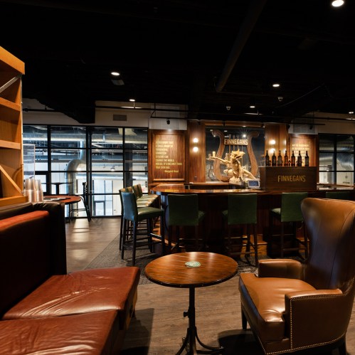 A cozy bar interior featuring wooden accents, leather seating, and a statue, with large windows in the background.