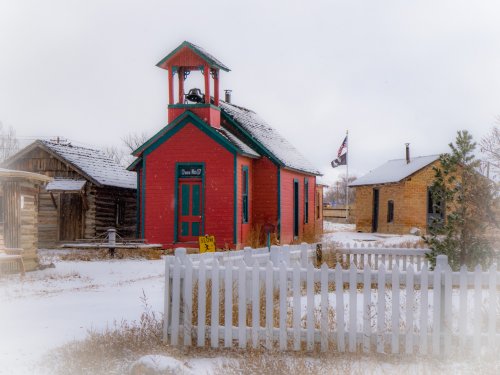 A colorful red schoolhouse with a bell tower, surrounded by snow and rustic buildings, under a cloudy sky.