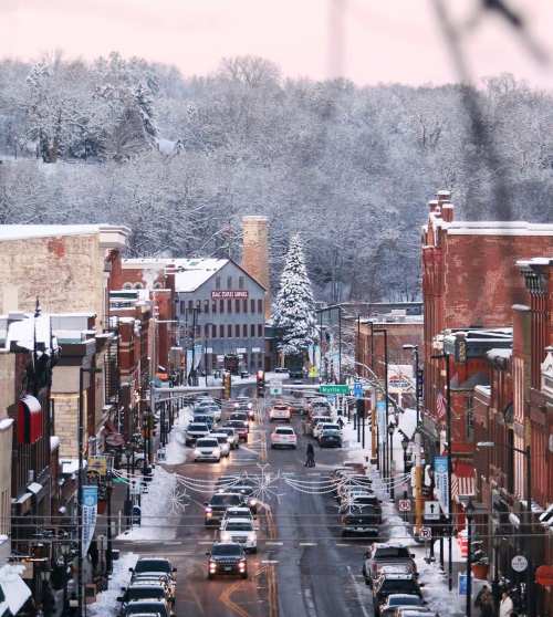 A snowy downtown street lined with historic buildings, cars, and a large Christmas tree in the distance.