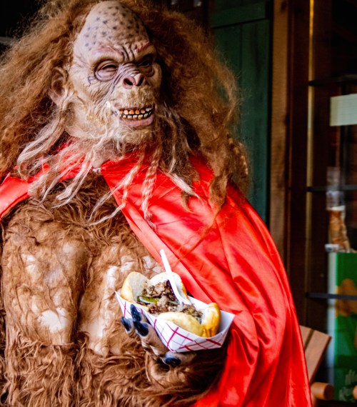 A creature with long hair and a red cape holds a sandwich, standing in a casual outdoor setting.