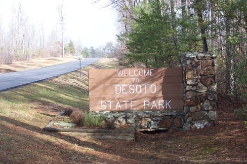 A wooden sign reading "Welcome to Desoto State Park" beside a road, surrounded by trees and natural scenery.