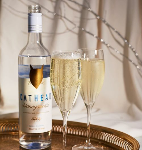 A bottle of Cathead Honeysuckle vodka with two fluted glasses of sparkling drink on a decorative tray.