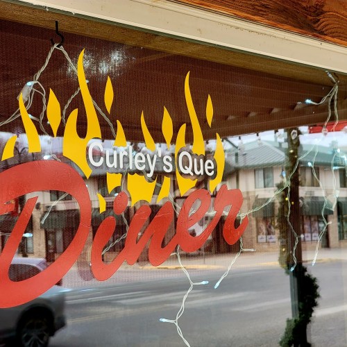 Sign for "Curley's Que Diner" with flame graphics and decorative lights in the window.