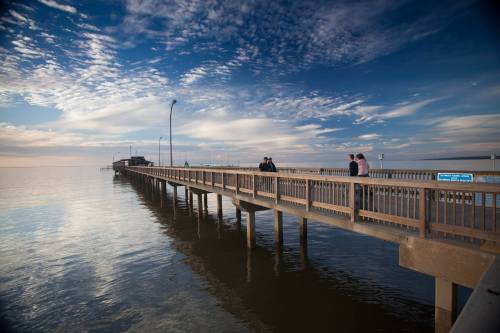 A wooden pier extends over calm water, with people walking and a scenic sky filled with clouds in the background.